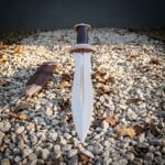 Hand forged roman pugio dagger with polished waisted midrib blade, engraved bronze guard and dark grip standing point down in pale gravel beside its leather sheath