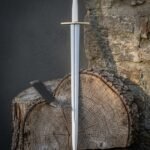 Hand forged medieval arming sword with polished tapering blade, brass wheel pommel and straight crossguard standing upright against a cut log and stone wall