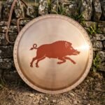 Hand made bronze faced hoplon shield, a Greek aspis about 30 inches across with a riveted rim, painted running boar device and leather carry strap against a dry stone wall