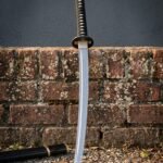 Hand forged damascus katana with a patterned folded steel blade and black ito wrapped tsuka leaning against a lichen covered brick wall above its lacquered black saya