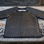 Blackened scale armor shirt of riveted steel scales on black leather with a runic throat plaque laid flat on a weathered wooden table
