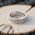 Hand made viking rune ring with a four strand Celtic knotwork band and Elder Futhark cut inside the shank, resting on a cut log beside a linen pouch