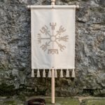 Hand made vegvisir banner in natural cotton canvas hung from a turned ash cross bar with cotton tassels, standing against a weathered stone wall