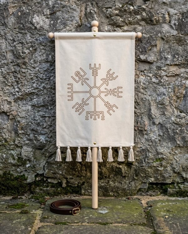 Hand made vegvisir banner in natural cotton canvas hung from a turned ash cross bar with cotton tassels, standing against a weathered stone wall