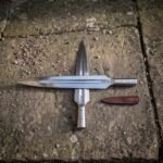 Two hand forged viking spearhead blades in satin carbon steel with rune cut sockets and knotwork panels beside a brown leather sheath on a stone slab.