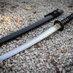 Hand forged wakizashi sword with a visible clay hardened hamon and engraved brass habaki lying on pale gravel beside its lacquered black saya among autumn leaves