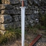 Hand forged valknut sword with a broad fullered polished blade and five lobed pommel standing against a dry stone wall beside a brown leather scabbard