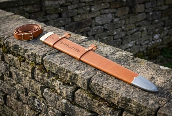 Tan leather sword scabbard tooled with Elder Futhark runes and fitted with a steel chape, laid on a mossy dry stone wall beside its matching buckled belt