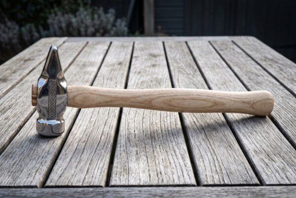 Hand forged 1095 steel cross pein hammer with a mirror polished head, engraved knotwork cheek and shaped hickory handle lying on a weathered wooden table