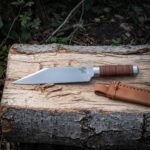 Hand forged viking seax with a broken back blade, etched raven and stacked leather handle lying on a split log beside its hand stitched tan leather sheath
