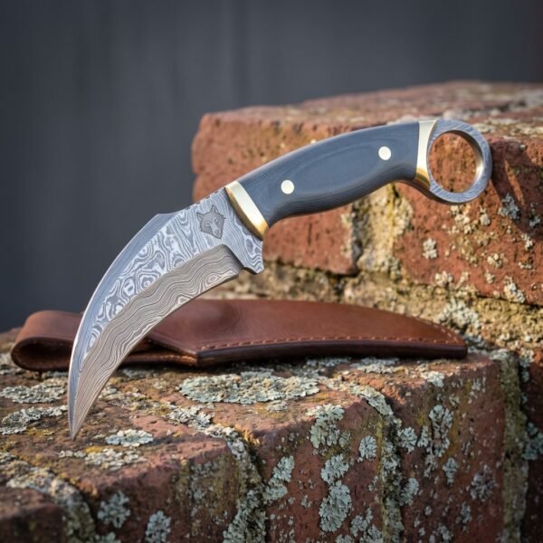 Hand forged damascus karambit knife with curved layered blade, green micarta scales, brass bolster and steel finger ring resting on a brick wall above its leather sheath