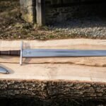 Hand forged medieval arming sword with a wheel pommel, long cross guard and fullered polished blade laid on a split oak slab beside a brown leather scabbard