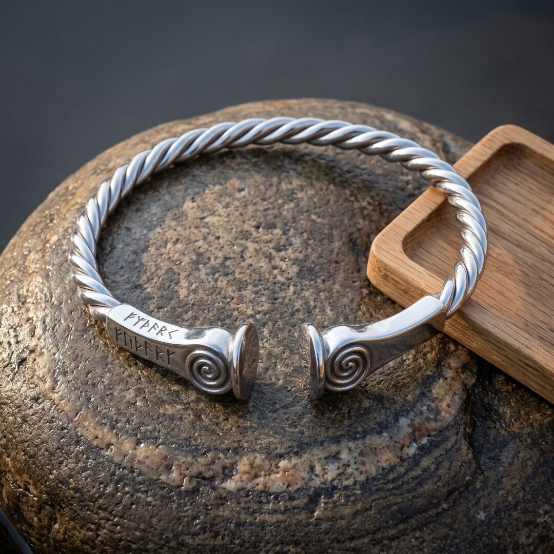 Gemini_Generated_Image_o25kapo25kapo25k Hand made viking arm ring with a twisted silver finish band, raised spiral terminals and Elder Futhark runes stamped near the ends, resting on weathered grey stone