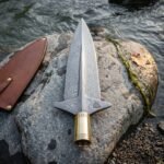 Hand forged damascus viking spearhead with winged lugs, valknut engraving and a runed brass socket resting on a river boulder beside a leather sheath