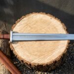 Hand forged rune engraved sword with Elder Futhark inscription cut into the fuller, russet leather grip and domed steel pommel lying on an oak log round
