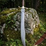 Hand forged damascus viking seax with a broken back blade, etched Valknut, rosewood grip and brass guard standing against a mossy boulder beside its tooled brown leather sheath