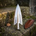 Mirror polished norse spearhead in 1095 steel with winged lugs, knotwork engraving and a runed socket standing on mossy stone beside a leather sheath
