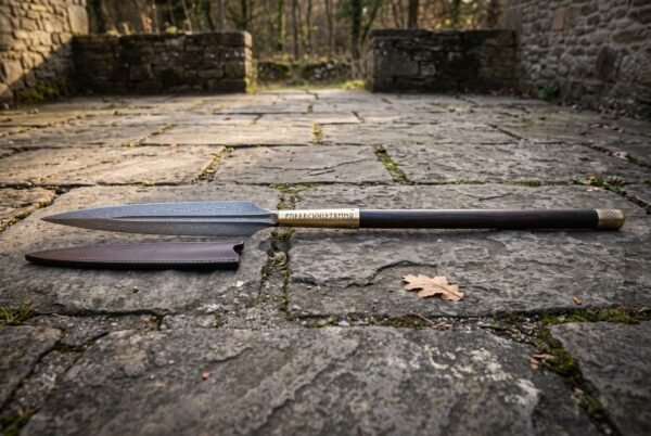 Hand forged Viking spear with a Damascus leaf blade in 1095 and 15N20, rune cut brass socket and dark stained ash shaft lying on flagstones beside its leather sheath