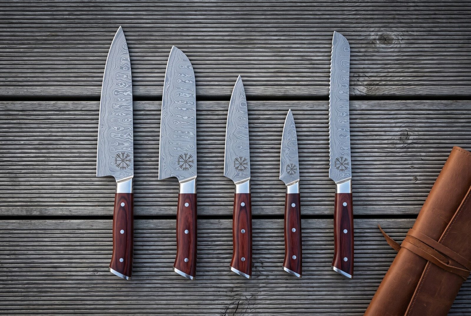 Gemini_Generated_Image_owsvxlowsvxlowsv Hand forged damascus chef knife set of five with rosewood handles and Vegvisir etched blades laid out on grey decking beside a rolled tan leather knife roll