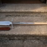 Hand forged boar spear with a broad polished leaf blade, curved steel crossbar and pale ash haft lying on stone steps beside a russet leather blade sheath