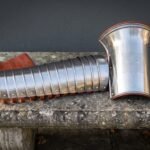 Polished steel manica arm guard of articulated bands beside a flared knotwork engraved bracer with russet leather straps on a stone bench outdoors.