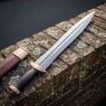 Hand forged damascus gladius short sword with layered blade, cut Greek key border and solid bronze pommel lying on a lichen covered brick wall beside its scabbard
