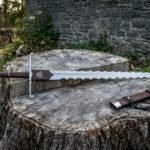 Hand forged arming sword with a flame bladed edge, scrolled crossguard and Valknut stamped leather rain guard lying on a tree stump beside its brown leather scabbard