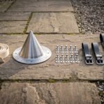 Hand raised conical shield boss in steel with an engraved Elder Futhark rune ring laid on flagstones beside leather rim binding, rivets and black leather straps