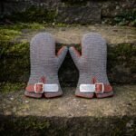 Pair of hand forged chainmail mittens in bright mild steel butted mail with russet leather cuffs and engraved knotwork buckle plates resting on a mossy stone wall