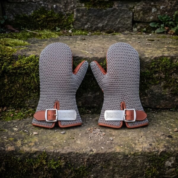 Pair of hand forged chainmail mittens in bright mild steel butted mail with russet leather cuffs and engraved knotwork buckle plates resting on a mossy stone wall