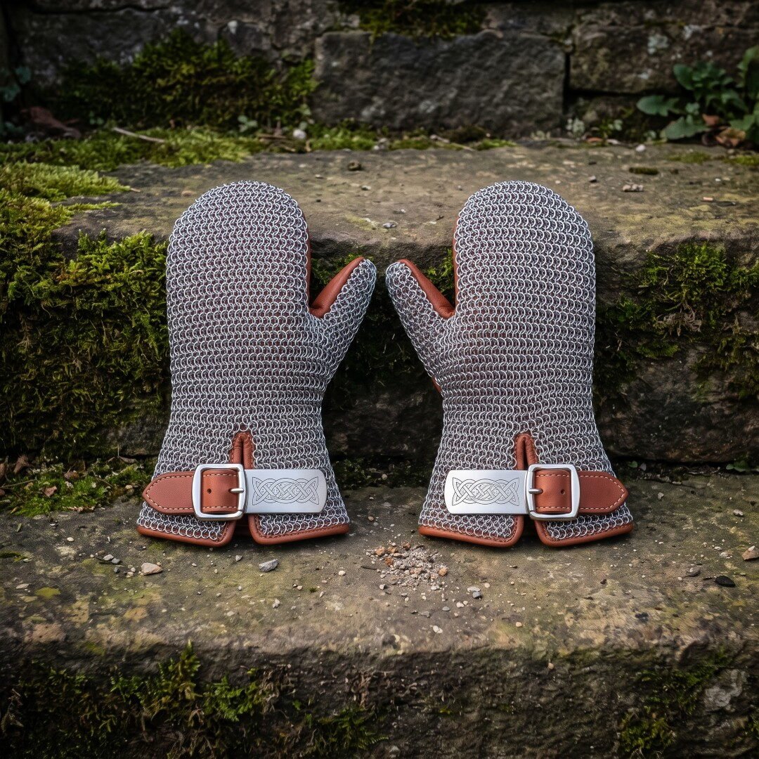 Gemini_Generated_Image_qcsdv2qcsdv2qcsd Pair of hand forged chainmail mittens in bright mild steel butted mail with russet leather cuffs and engraved knotwork buckle plates resting on a mossy stone wall