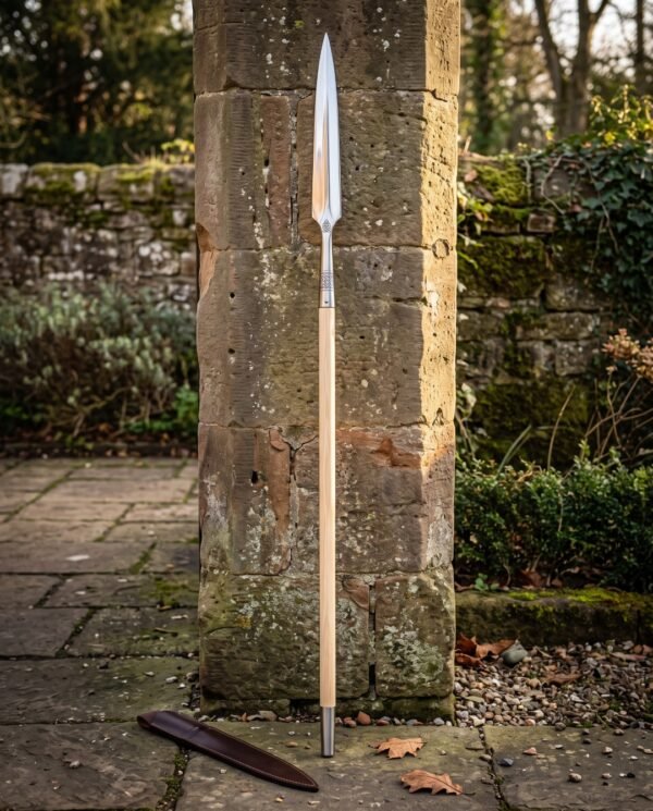 Hand forged viking spear standing against a stone pillar, long steel head with a knotwork cut socket on an ash shaft with a leather blade cover below.