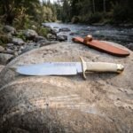 Hand forged viking bowie knife with polished clip point blade, rune engraving, pale bone handle and brass guard resting on a river boulder beside its leather sheath