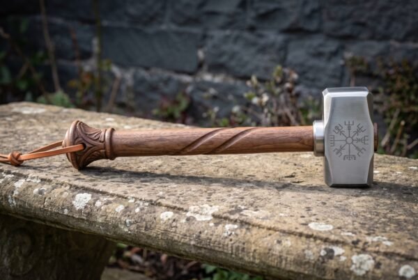Hand forged blacksmith hammer with a polished 1095 steel head engraved with the Vegvisir and a carved spiral hardwood handle, lying on a weathered stone bench
