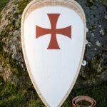 Templar shield of kite form in planked wood, a red cross pattee on a white field with a knotwork border band, leaning on a mossy boulder beside a leather belt.