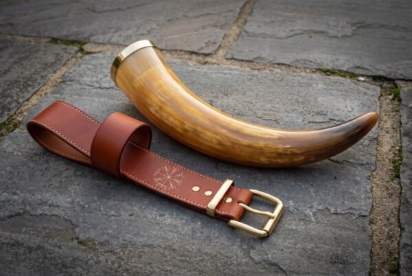 Hand made viking drinking horn with a polished brass rim resting on grey flagstone beside a tan leather belt hanger stamped with a Vegvisir runic compass.
