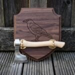 Hand forged bearded axe with a light ash haft and brass brackets mounted on a walnut viking axe wall plaque engraved with a standing raven, shown on weathered decking.