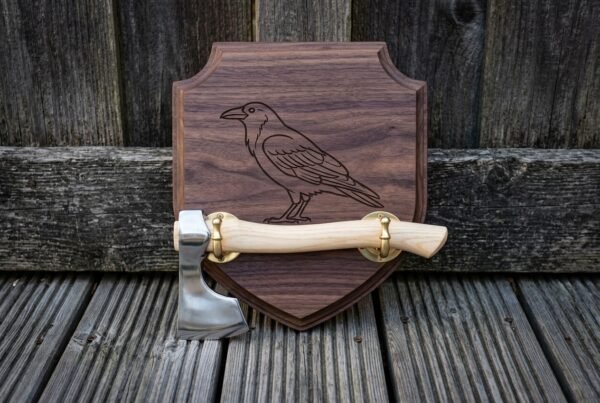 Hand forged bearded axe with a light ash haft and brass brackets mounted on a walnut viking axe wall plaque engraved with a standing raven, shown on weathered decking.