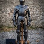 Blackened hand forged plate armour suit on a wooden stand against a stone wall, showing fluted breastplate, engraved runes, articulated pauldrons, gauntlets and sabatons.