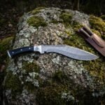 Hand forged damascus kukri with a flowing pattern welded blade, engraved Valknut and buffalo horn handle, lying on a lichen covered rock beside its leather sheath