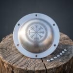 Polished steel Vegvisir shield boss with the stave inside a knotwork ring, eight drilled flange holes, on a cut tree stump beside a row of dome rivets.