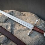 Hand forged arming sword with a polished fullered blade, knotwork engraved crossguard, leather wrapped grip and disc pommel, lying on a boulder beside its brown leather scabbard.