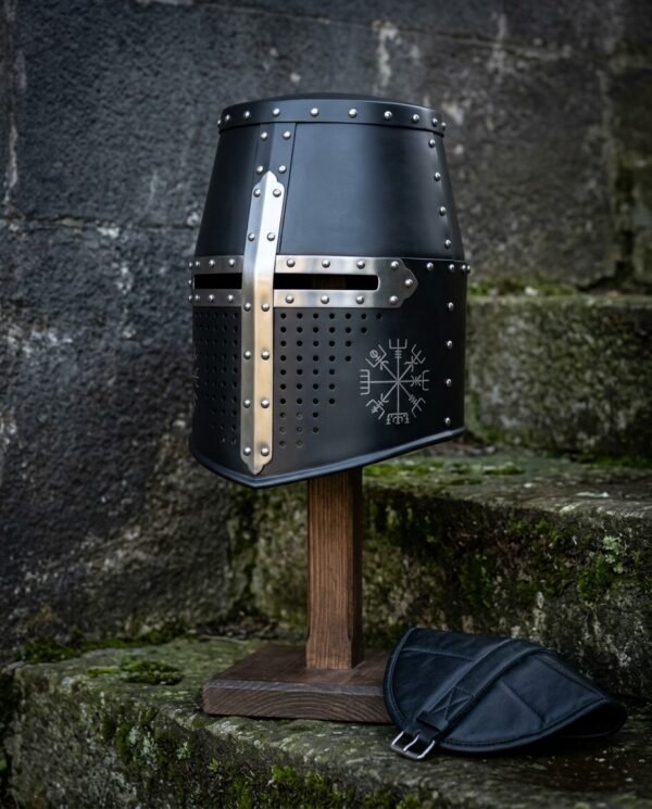 Blackened crusader helmet, a hand forged 16 gauge great helm with a polished steel cross reinforce, domed rivets and a chased Vegvisir, on a wooden stand by mossy steps