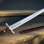 Hand forged knightly sword with a polished fullered blade, engraved knotwork panel, brass crossguard and pommel and oxblood leather grip, resting on a stone step beside its burgundy scabbard.