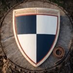 Heater shield in planked wood quartered navy blue and white with a red border and cut knotwork band, lying on a cut tree stump beside a coiled leather strap.