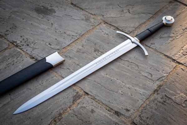 Medieval arming sword in polished high carbon steel with curved crossguard, black leather grip and wheel pommel, lying on flagstones beside a black leather scabbard
