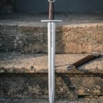 Damascus viking sword with Elder Futhark runes cut into the broad blade, leather wrapped grip and wheel pommel, standing on stone steps beside a timber scabbard