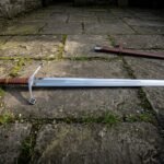 Hand forged medieval longsword with a long tapering fullered blade, curved crossguard with ring terminals and a two hand leather grip, lying on flagstones beside its leather scabbard.