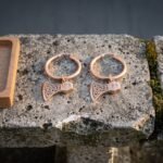Hand made celtic earrings as rose tone hoops with bearded Viking axe head charms and cut knotwork panels laid on a weathered grey stone slab
