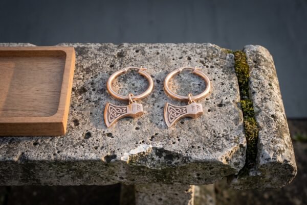 Hand made celtic earrings shown as rose tone hoops with bearded Viking axe head charms and cut knotwork panels laid on a weathered grey stone slab beside a small walnut tray